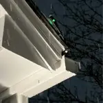 Close-up of a white house eaves with a small green LED light on a detector at dusk, bare branches in the background.